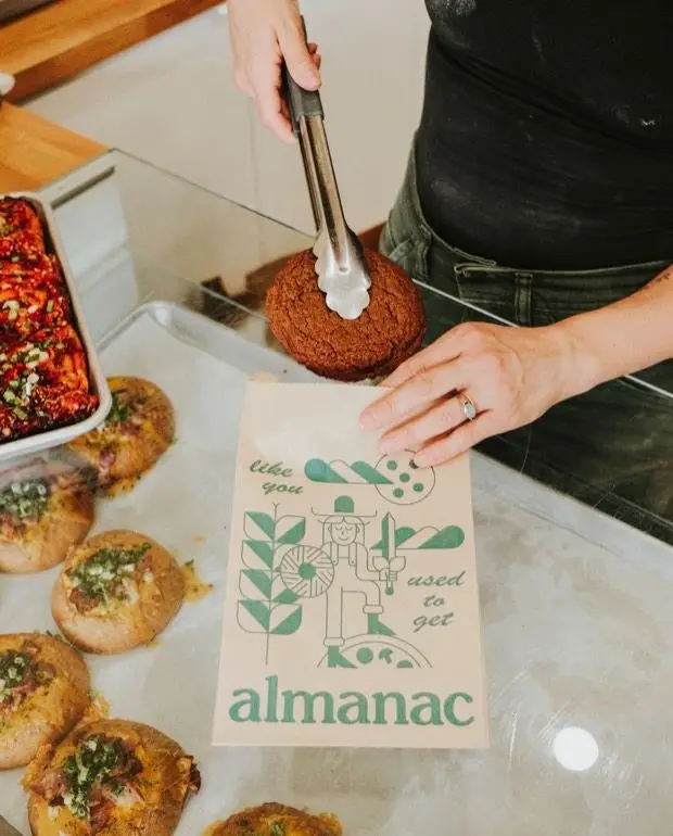 A baker bagging a pastry at the Almanac counter, beside a tray of savoury buns.
